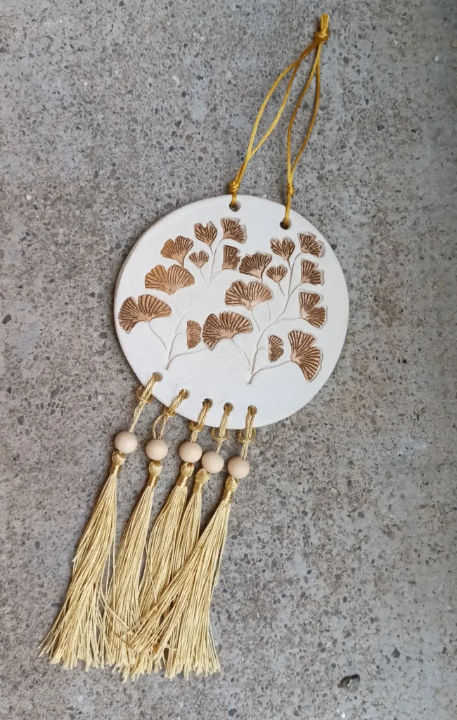 Suspension en grès blanc, motif feuilles de ginkgo biloba, avec ficelle et pompons dorés, perles en bois, diamètre 10,5 cm, Isabelle Morales